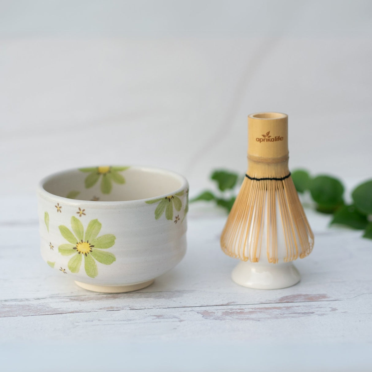 The Green Flowers Ceramic Matcha Bowl, adorned with green flowers and paired with a bamboo whisk, sits elegantly on a white surface surrounded by lush leaves. Handcrafted using Minoyaki pottery techniques, this teaware beautifully captures the essence of tradition and nature.