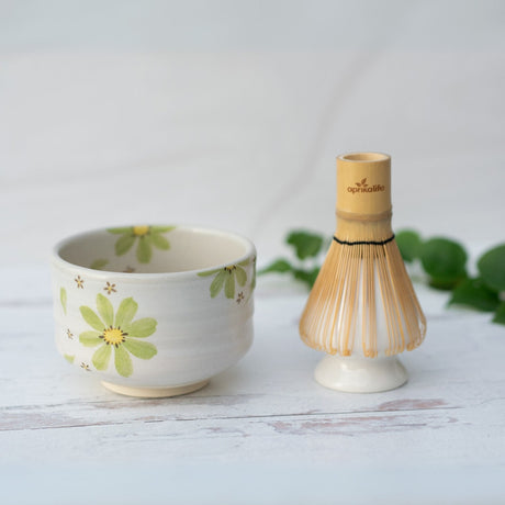 The Green Flowers Ceramic Matcha Bowl, adorned with green flowers and paired with a bamboo whisk, sits elegantly on a white surface surrounded by lush leaves. Handcrafted using Minoyaki pottery techniques, this teaware beautifully captures the essence of tradition and nature.