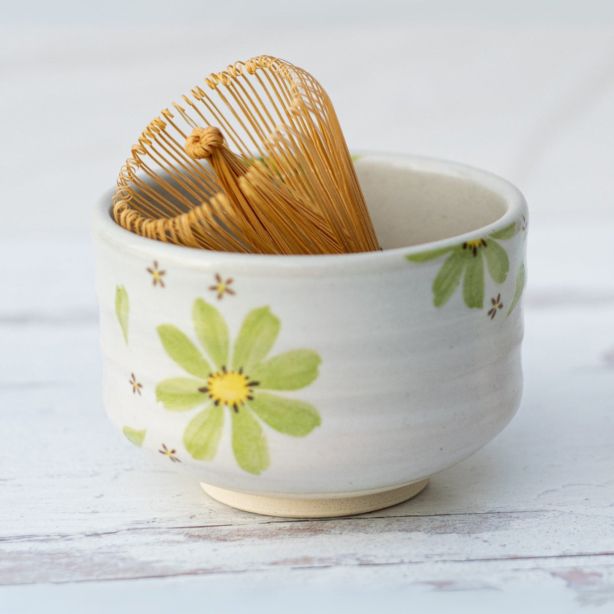Showcasing traditional Minoyaki pottery, the Green Flowers Ceramic Matcha Bowl features a green flower design and holds a tea whisk—an essential piece for any handcrafted matcha teaware collection on a pristine white surface.