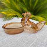 Glass hen-shaped container on a textured surface with green foliage in the background