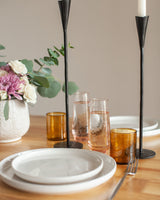 Dining table setting with white plates, amber glasses, black candle holders, and a vase with flowers.