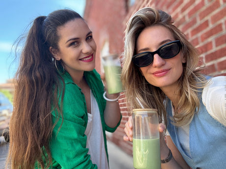 Two women smile outdoors, holding clear cups of matcha lattes made with Ceremonial Grade Matcha Latte Powder Mix with Natural Vanilla Flavor, standing in front of a brick wall.