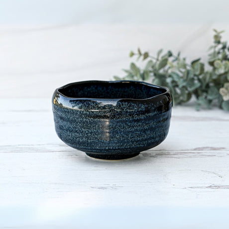 The Indigo Glaze Matcha Bowl, a dark blue Minoyaki ceramic piece with a textured surface, rests beautifully on a light wooden table with greenery behind it, embodying the artistry of Japan's handcrafted matcha tea bowls.