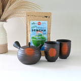 A Japanese Kyusu Set, including a 270ml teapot with filters and two teacups, rests on a pristine white surface alongside organic sencha tea, with a delicate vase adding elegance to the background.