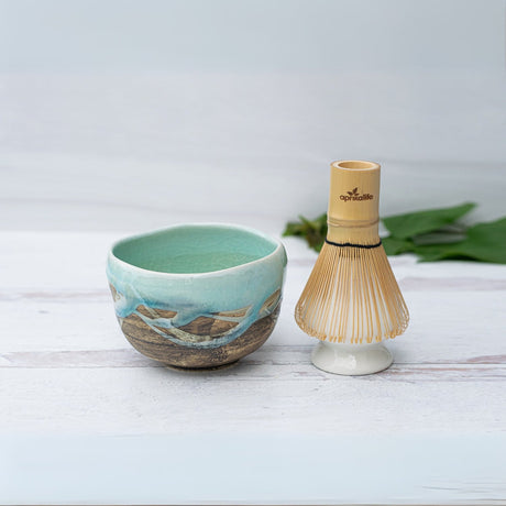 The Japanese Matcha Tea Bowl Blue & Brown Glaze, Bamboo Matcha Whisk and Whisk Holder Set rests on a light surface with green leaves behind—crafted using traditional Minoyaki pottery techniques.
