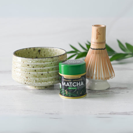 The Kaneni set includes a white ceramic matcha bowl, bamboo whisk, whisk holder, and a tin of organic Japanese matcha on a light wooden surface.
