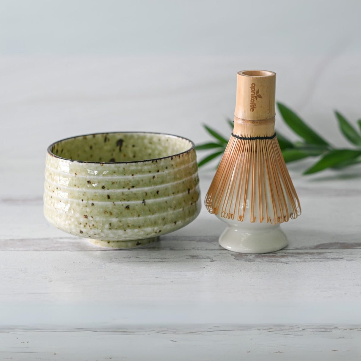 The Kaneni White Ceramic Matcha Bowl and Bamboo Matcha Whisk from the Organic Japanese Matcha Set, crafted with Minoyaki techniques, rest elegantly on a light wood surface with green leaves in the background.