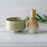 The Kaneni White Ceramic Matcha Bowl and Bamboo Matcha Whisk from the Organic Japanese Matcha Set, crafted with Minoyaki techniques, rest elegantly on a light wood surface with green leaves in the background.