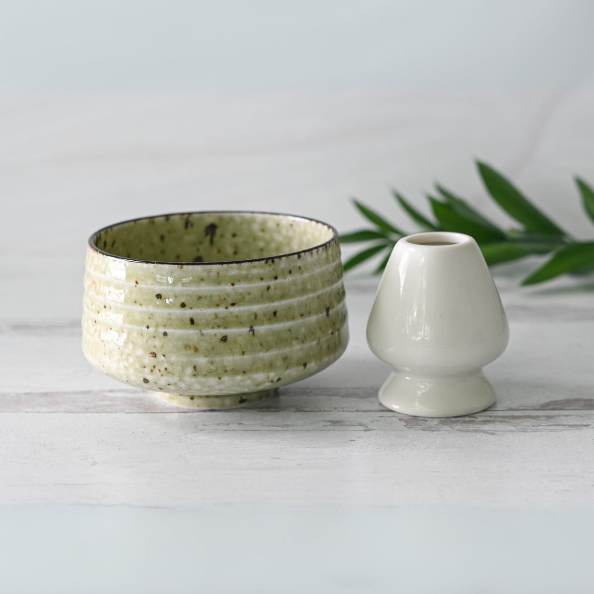 A Kaneni White Ceramic Matcha Bowl, crafted using Minoyaki techniques, and a small white vase sit on a light surface adorned with green leaves.