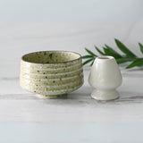 A Kaneni White Ceramic Matcha Bowl, crafted using Minoyaki techniques, and a small white vase sit on a light surface adorned with green leaves.