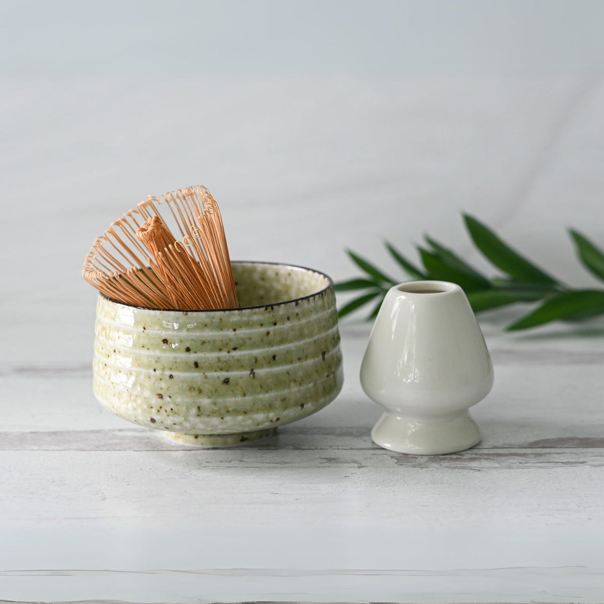 The Kaneni Matcha Set includes a white ceramic bowl crafted with Minoyaki techniques, a bamboo whisk, and a holder on a light surface, surrounded by green leaves—a quintessential example of Japanese pottery.