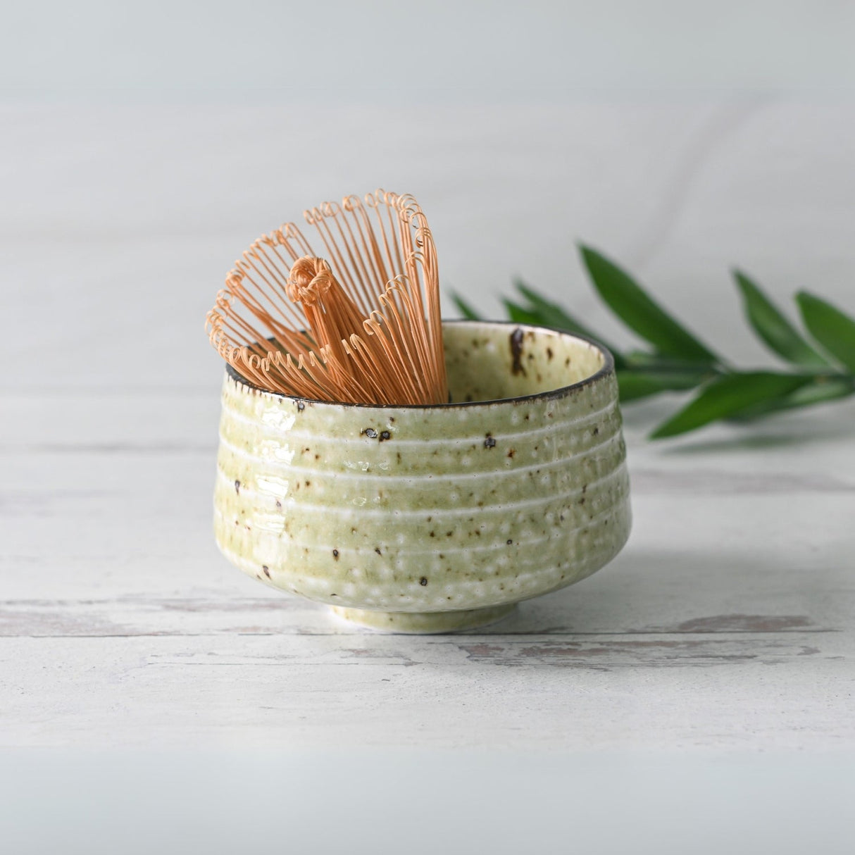 The Kaneni White Ceramic Matcha Bowl rests alongside a bamboo matcha whisk, nestled on its holder, part of the Organic Japanese Matcha Set, with a green leaf nearby on the white wooden surface.