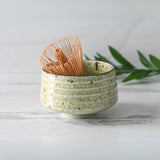 The Kaneni White Ceramic Matcha Bowl rests alongside a bamboo matcha whisk, nestled on its holder, part of the Organic Japanese Matcha Set, with a green leaf nearby on the white wooden surface.