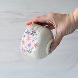 A hand cradles a Kohiki Pink Sakura Ceramic Matcha Bowl, adorned with a pink and purple floral design, reminiscent of those used in a traditional matcha tea ceremony.