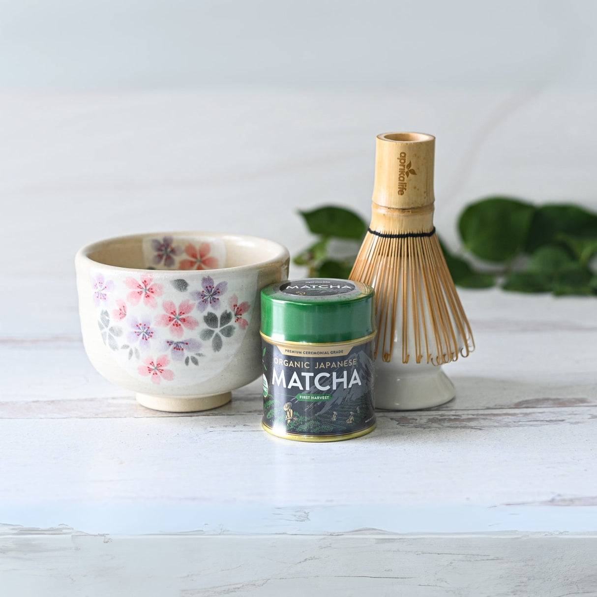 A white surface displays a traditional matcha tea ceremony set, featuring the Kohiki Pink Sakura Ceramic Matcha Bowl alongside a floral teacup, tin of matcha powder, and bamboo whisk.
