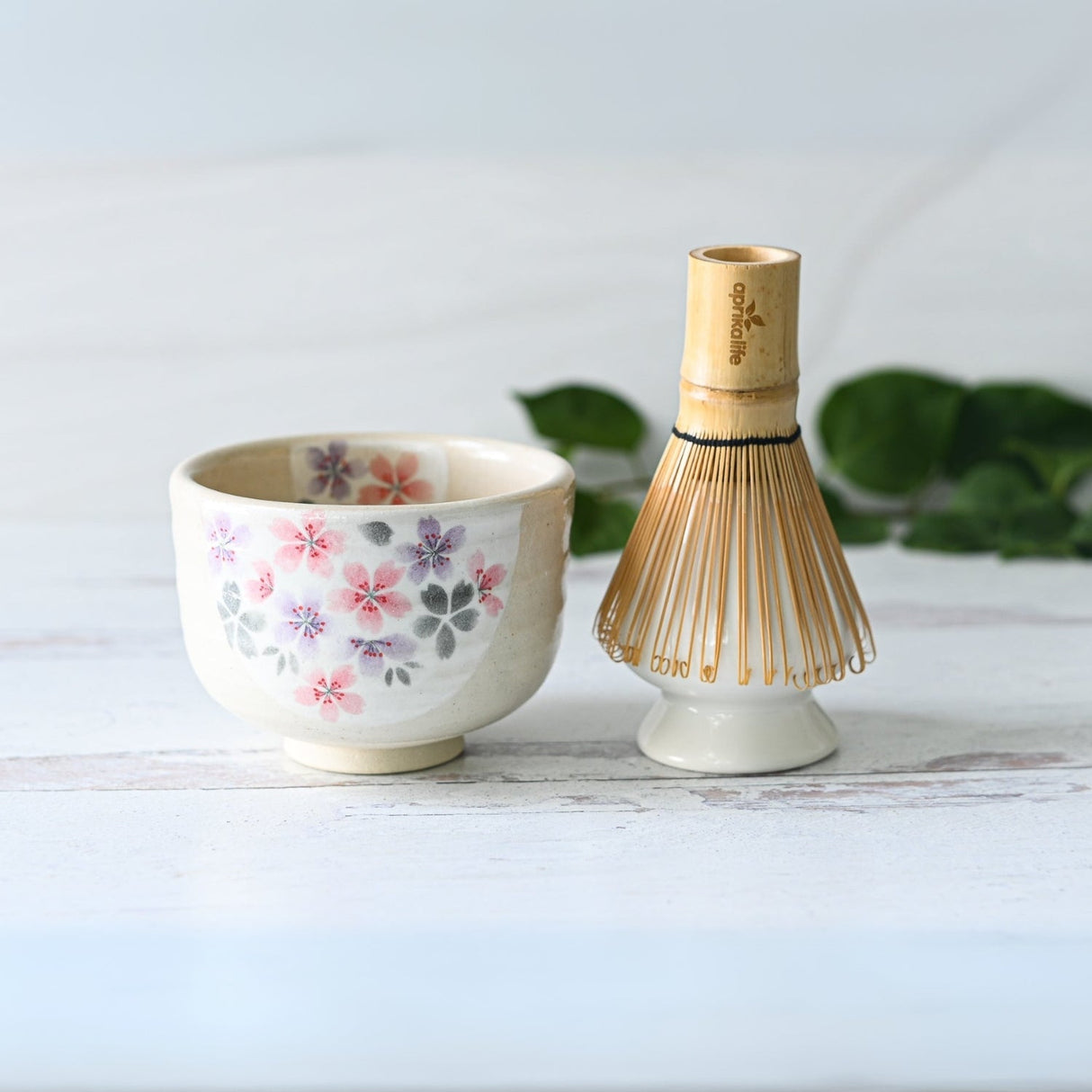 The Kohiki Pink Sakura Ceramic Matcha Bowl, featuring a floral design and accompanied by a bamboo whisk, sits on a light wooden surface among green leaves, capturing the spirit of a traditional Matcha tea ceremony.