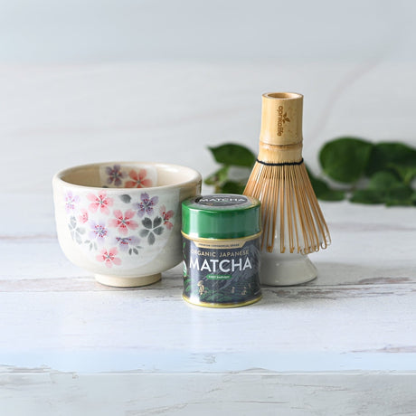 A white surface displays a traditional matcha tea ceremony set, featuring the Kohiki Pink Sakura Ceramic Matcha Bowl alongside a floral teacup, tin of matcha powder, and bamboo whisk.