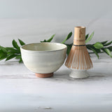The Kohiki White Clay Matcha Bowl and bamboo whisk sit elegantly on a white surface, surrounded by lush green leaves, creating a serene setting for your next matcha tea ritual.