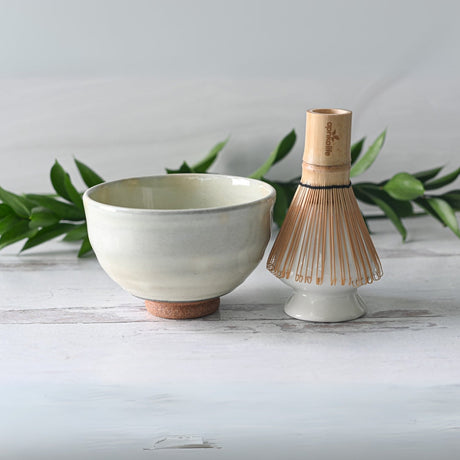 The Kohiki White Clay Matcha Bowl and bamboo whisk sit elegantly on a white surface, surrounded by lush green leaves, creating a serene setting for your next matcha tea ritual.