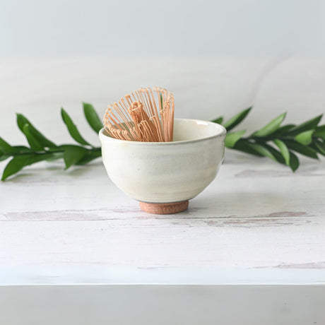 The Kohiki White Clay Matcha Bowl, featuring a whisk inside, sits on a white surface with green leaves in the background.