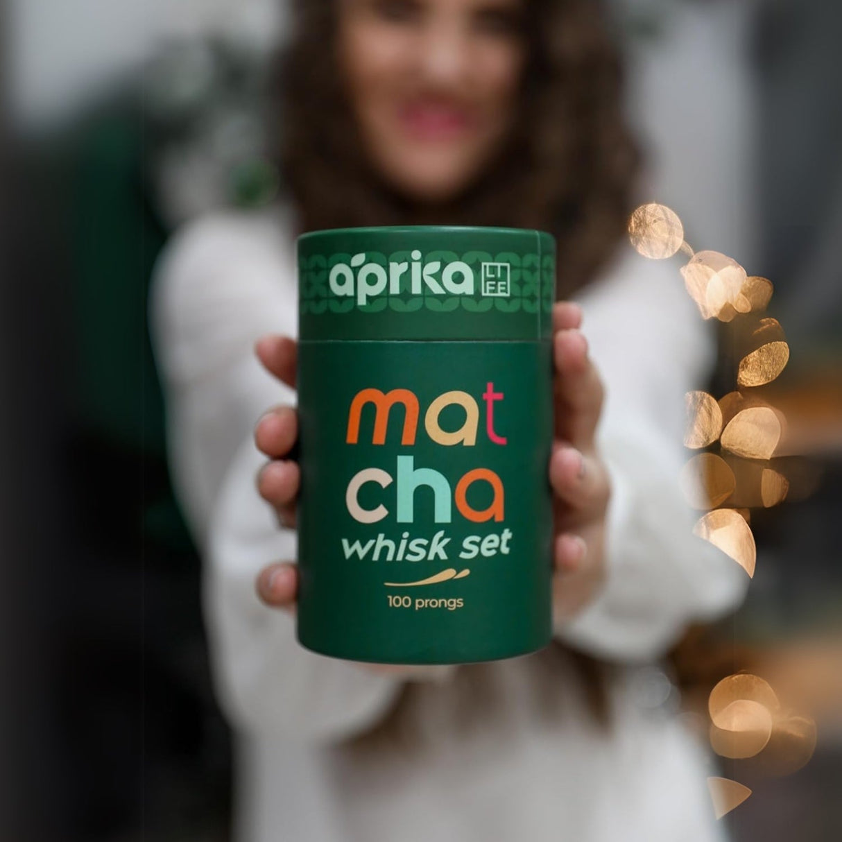 A person holds a Matcha Bamboo Whisk Set container while bokeh lights twinkle on the right, creating a serene matcha experience.