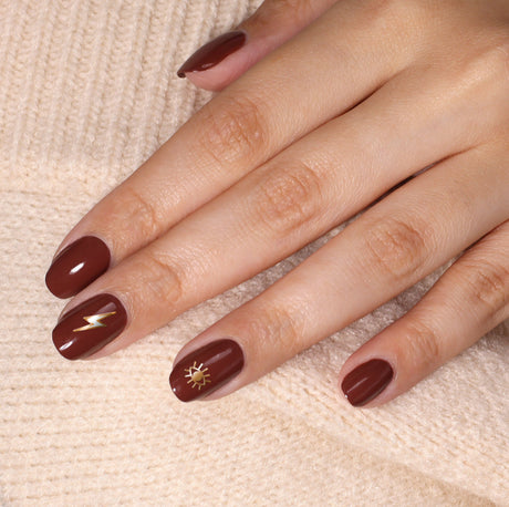 A hand with neatly manicured nails painted in brown, adorned with LONDONTOWN's Nail Decals - Cosmic featuring a lightning bolt on the ring finger and a sun symbol on the middle finger. The person is wearing a beige sweater.