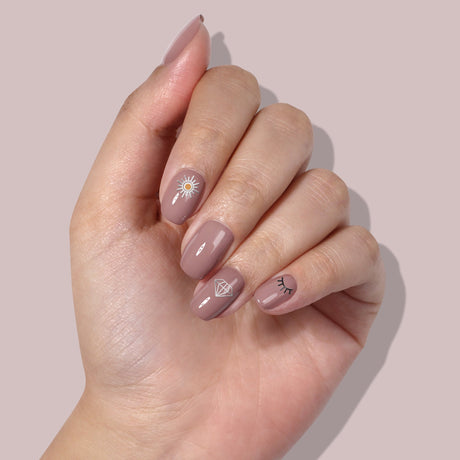 A hand with manicured nails painted in a nude shade features small sun, diamond, and eyelash designs using LONDONTOWN's Nail Decals - Cosmic, showcasing easy nail art at its finest.