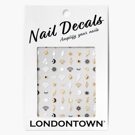 A package of LONDONTOWN Nail Decals - Cosmic featuring various designs like sun, moon, lightning bolts, diamonds, stars, and eyes in gold and black on a white background. The text says "Nail Decals - Amplify your nails with easy nail art.