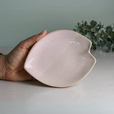 A hand holds a pink, leaf-shaped ceramic dish against green foliage, reminiscent of a Pink LOVE Ceramic Tea Cup with Saucer.