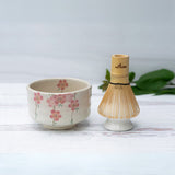 On a white surface lies the Pink Sakura Flowers Ceramic Matcha Bowl with a bamboo matcha whisk, part of an exquisite set including a whisk holder and organic Japanese matcha.
