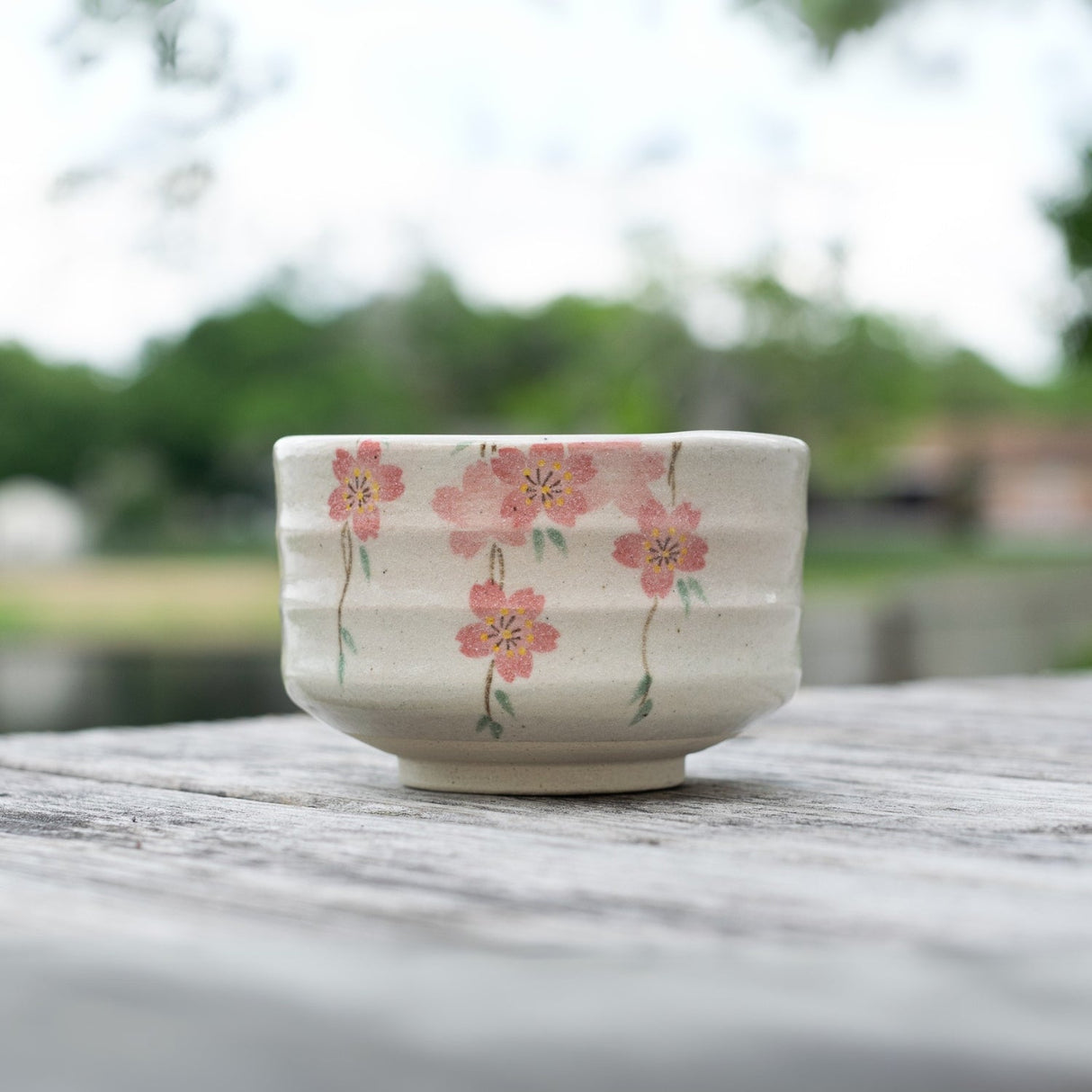The Pink Sakura Flowers Ceramic Matcha Bowl from the Japanese pottery matcha accessory set is elegantly placed on a wooden table with blurred greenery in the background.