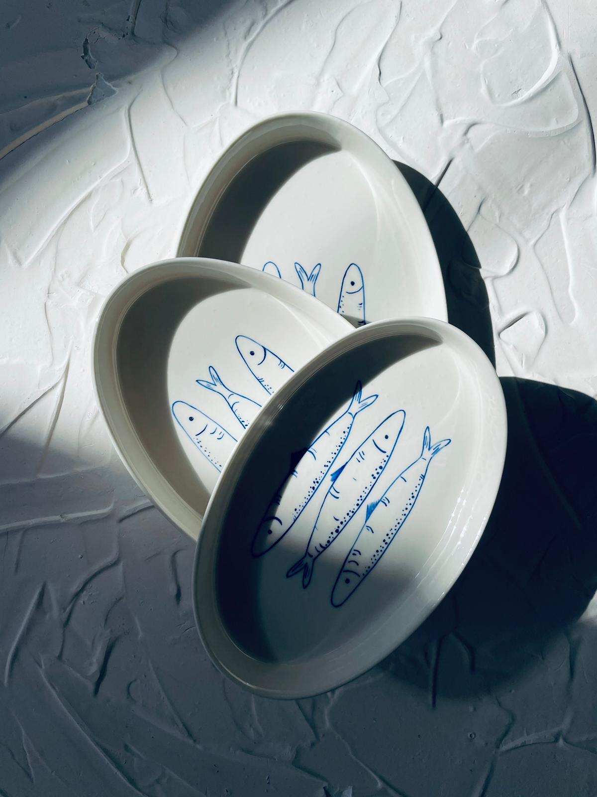 White ceramic plates with blue fish designs on a textured white surface. Sardines Nesting Plates