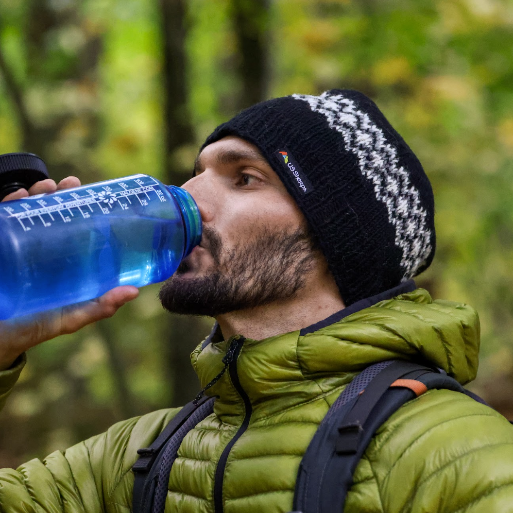 Man in green jacket and black beanie drinking from a blue water bottle outdoors.