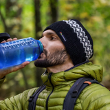 Man in green jacket and black beanie drinking from a blue water bottle outdoors.