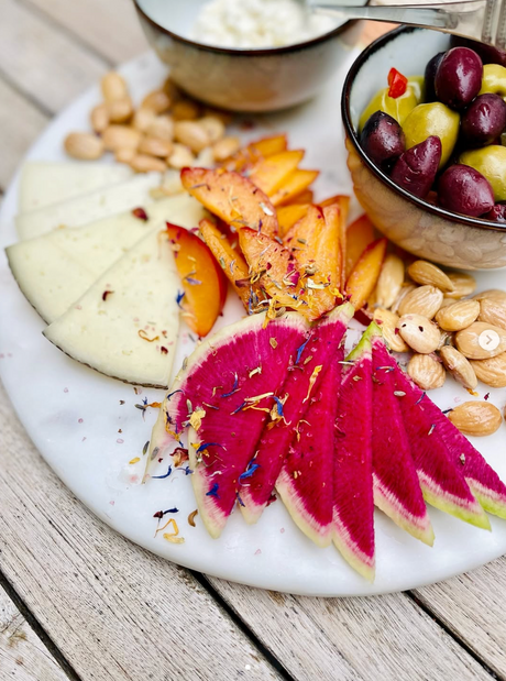 Platter with sliced vegetables, nuts, and a bowl of olives on a wooden surface