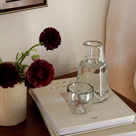 Clear glass carafe and candle on a book with a vase of dark red flowers on a wooden surface.