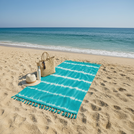 Turquoise striped beach towel on sand with a straw bag, hat, and bottle.