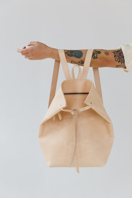 Beige backpack held by a person with tattoos on a light gray background. The Folded Backpack - Natural