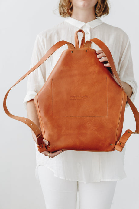 The Folded Backpack - Saddle. Person holding a brown leather backpack against a white background