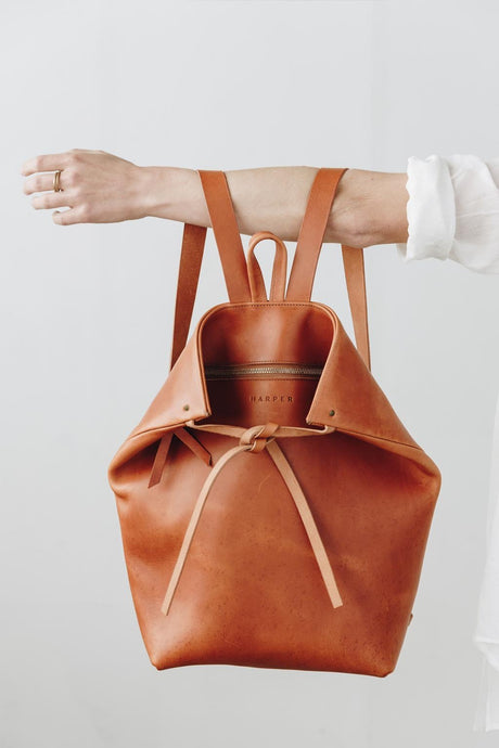 The Folded Backpack - Saddle. Brown leather backpack held by a person on a light gray background