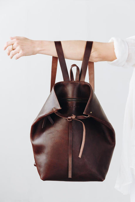 The Folded Backpack - Walnut. Brown leather backpack held by a person on a white background