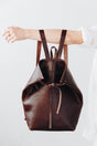 The Folded Backpack - Walnut. Brown leather backpack held by a person on a white background