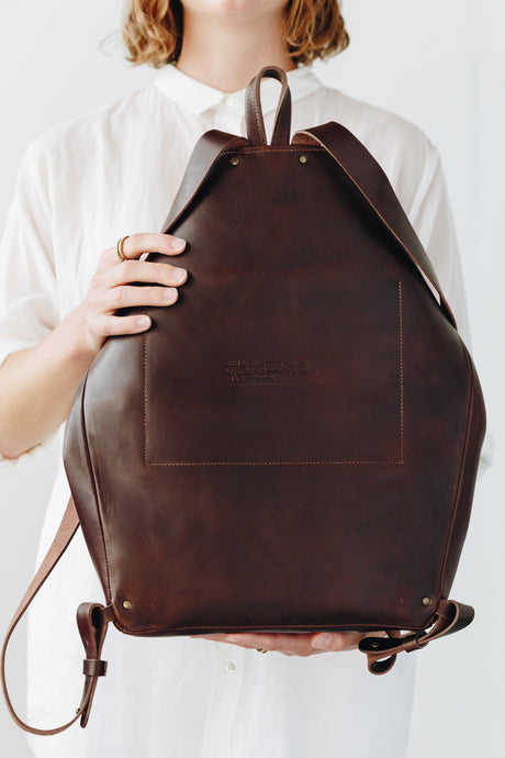 The Folded Backpack - Walnut. Brown leather backpack held by a person against a white background