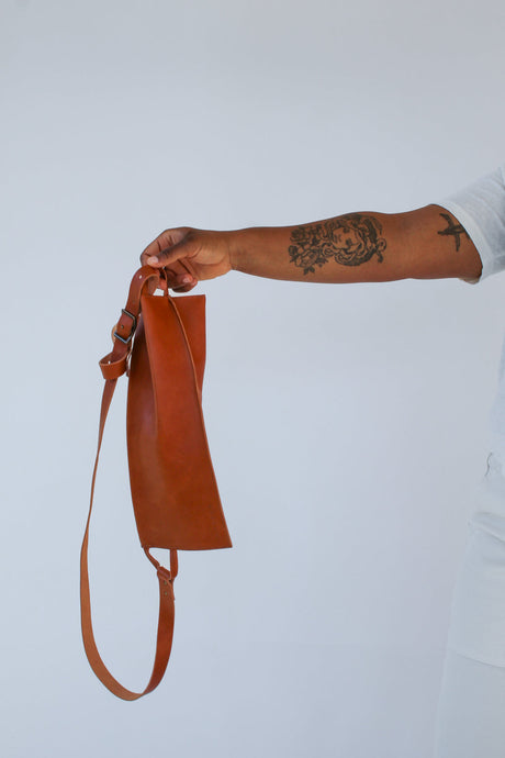 The Offset Sling Bag - Saddle. Brown leather pouch held by a person with tattoos on a light gray background. 