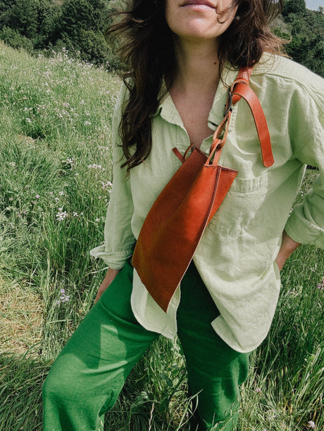 The Offset Sling Bag - Saddle. Person wearing a light green shirt and green pants with an orange leather bag in a grassy field.