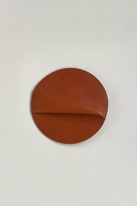Brown leather round object on a light gray background. The Pleated Clutch - Saddle