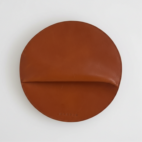 Brown leather round object on a light gray background. The Pleated Clutch - Saddle