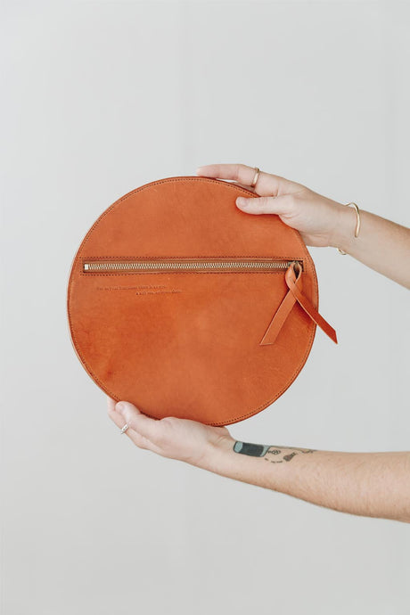 Round orange clutch bag held by a person against a light background. The Pleated Clutch - Saddle