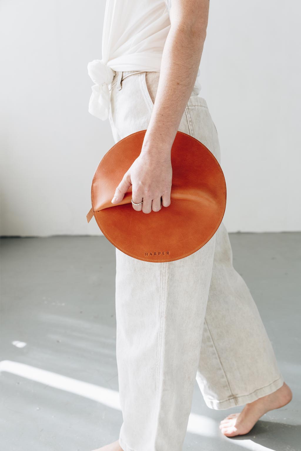 Person holding a round, brown leather clutch against a plain background. The Pleated Clutch - Saddle