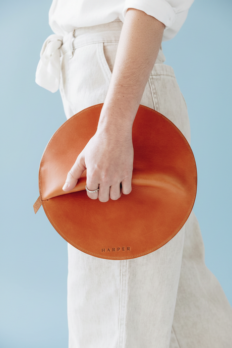 Person holding a round, brown leather clutch with 'Harper' branding against a light blue background. The Pleated Clutch - Saddle
